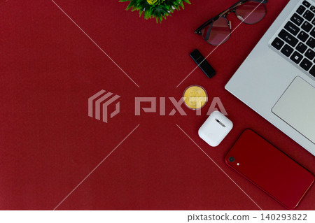Red surface holds tech and accessories. Laptop, phone, earbuds, and sunglasses rest nearby. Small plant adds green contrast to bold red. Cozy, stylish, modern scene for digital lifestyles 140293822