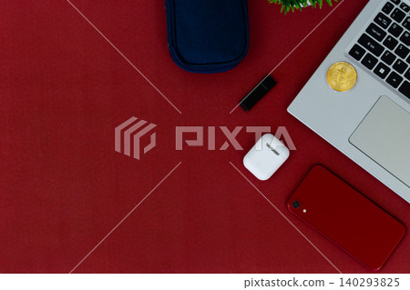 Red surface holds tech and accessories. Laptop, phone, earbuds, and sunglasses rest nearby. Small plant adds green contrast to bold red. Cozy, stylish, modern scene for digital lifestyles 140293825