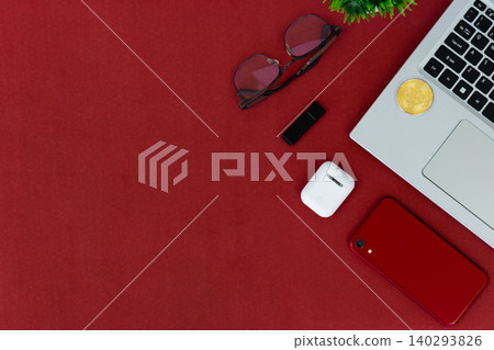 Red surface holds tech and accessories. Laptop, phone, earbuds, and sunglasses rest nearby. Small plant adds green contrast to bold red. Cozy, stylish, modern scene for digital lifestyles 140293826