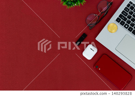 Red surface holds tech and accessories. Laptop, phone, earbuds, and sunglasses rest nearby. Small plant adds green contrast to bold red. Cozy, stylish, modern scene for digital lifestyles 140293828