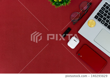 Red surface holds tech and accessories. Laptop, phone, earbuds, and sunglasses rest nearby. Small plant adds green contrast to bold red. Cozy, stylish, modern scene for digital lifestyles 140293829
