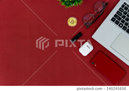 Red surface holds tech and style items. Laptop, phone, earbuds, and glasses create modern vibe. Golden coin adds digital finance touch. Minimalist layout invites creative use 140293830
