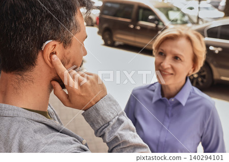 Adult man and woman communicate in a lively city setting, male using a hearing aid to enhance conversation amidst urban noise Adult man and woman communicate in a lively city setting, male using a hearing aid to enhance conversation amidst urban noise 140294101