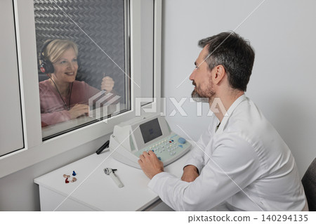 Hearing testing. Mature female patient wearing audiology headphones pressing button of response while audiometric testing in soundproof audiology booth Hearing testing. Mature female patient wearing audiology headphones pressing button of response while audiometric testing in soundproof audiology booth 140294135