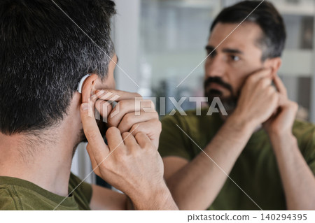 Adult man explores the comfort and fit of a new behind-the-ear hearing aid, adjusting it carefully in front of a mirror. Stylish hearing device 140294395