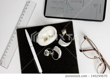 Top view showcases a hearing aid set alongside an ear model, ruler, glasses, and a writing pen on a dark surface. Hearing solutions 140294670