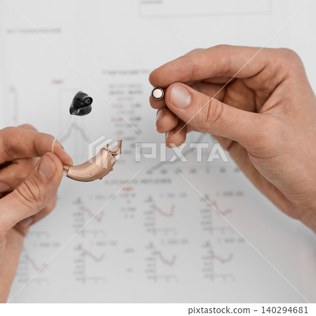 Two hands replacing a hearing aid battery surrounded by medical cards. Hearing aid maintenance services Two hands replacing a hearing aid battery surrounded by medical cards. Hearing aid maintenance services 140294681