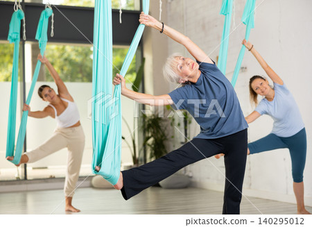 Senior woman practicing aerial yoga asana during group class in gym Senior woman practicing aerial yoga asana during group class in gym 140295012