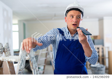 Worker in a protective helmet and blue overall discussing repair plan on the speakerphone of mobile phone Worker in a protective helmet and blue overall discussing repair plan on the speakerphone of mobile phone 140296099