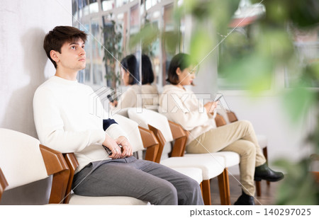 Young man browsing websites on smartphone while sitting in lobby of job center or employment office, waiting for interview or conversation with career counselor Young man browsing websites on smartphone while sitting in lobby of job center or employment office, waiting for interview or conversation with career counselor 140297025