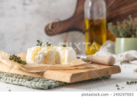 A rustic scene of fresh white ricotta cheese topped with golden honey and thyme sprigs resting on parchment paper and a wooden board with olive oil and kitchen tools in the soft blurred background 140297083