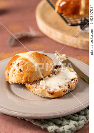 Vertical close-up of a sliced hot cross bun spread with melting butter on a grey plate with a gold knife in a rustic spring kitchen setting 140297084