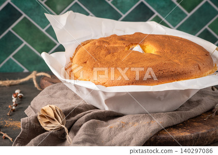 A golden Pao de Lo sponge cake wrapped in white parchment paper sits on a rustic wooden board against a green tiled wall with cozy kitchen decor and seasonal accents 140297086