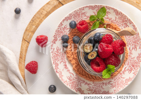 A close-up overhead shot features a glass jar of overnight oats with chia seeds topped with fresh raspberries and blueberries on a floral plate with a wooden spoon and mint garnish A close-up overhead shot features a glass jar of overnight oats with chia seeds topped with fresh raspberries and blueberries on a floral plate with a wooden spoon and mint garnish 140297088