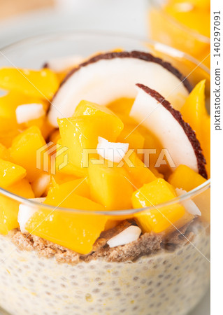 Creamy chia seed pudding in a glass topped with vibrant mango cubes and fresh coconut slices for a healthy breakfast or tropical vegan dessert served on a neutral aesthetic background with no marks 140297091
