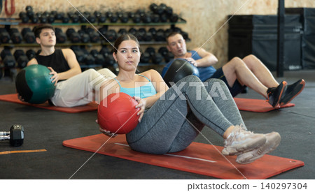 Strong young woman performing exercise for abs core with medicine ball on yoga mat during group workout in CrossFit gym center Strong young woman performing exercise for abs core with medicine ball on yoga mat during group workout in CrossFit gym center 140297304