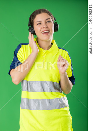 Female worker in reflective polo shirt enjoys listening to music on headphones and dancing on green background Female worker in reflective polo shirt enjoys listening to music on headphones and dancing on green background 140298211