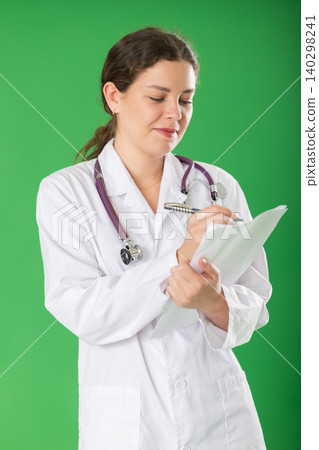 Young woman doctor in white coat with a stethoscope conducts a survey remotely green background Young woman doctor in white coat with a stethoscope conducts a survey remotely green background 140298241