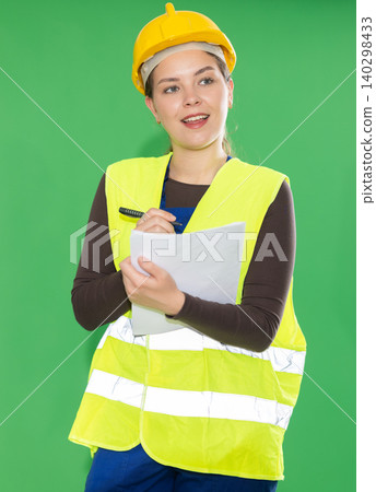 Portrait of girl worker dressed in reflective vest and hard hat with papers in hands checking work process standing against green screen Portrait of girl worker dressed in reflective vest and hard hat with papers in hands checking work process standing against green screen 140298433
