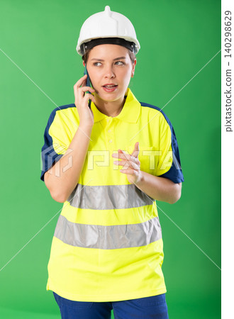 Young emotional girl wearing reflective polo and hard hat talking expressively to client by smartphone standing against green screen Young emotional girl wearing reflective polo and hard hat talking expressively to client by smartphone standing against green screen 140298629