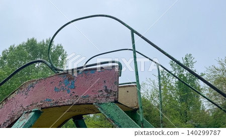 Old rusty metal playground slide with peeling paint against green trees Old rusty metal playground slide with peeling paint against green trees 140299787