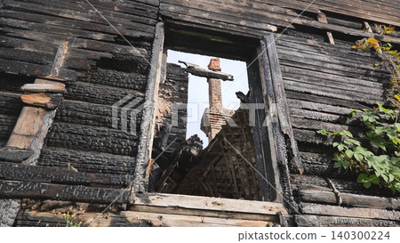 Burnt wooden house wall with an empty window frame and a distant brick chimney Burnt wooden house wall with an empty window frame and a distant brick chimney 140300224