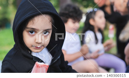 Young girl enjoying a refreshing popsicle outdoors with friends, her face messy Young girl enjoying a refreshing popsicle outdoors with friends, her face messy 140300315