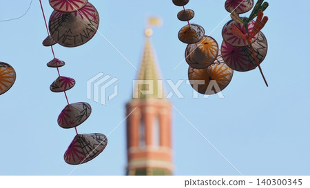 Traditional Vietnamese conical hat decorations hanging at an outdoor festival with a blurred historic tower in the background. Traditional Vietnamese conical hat decorations hanging at an outdoor festival with a blurred historic tower in the background. 140300345