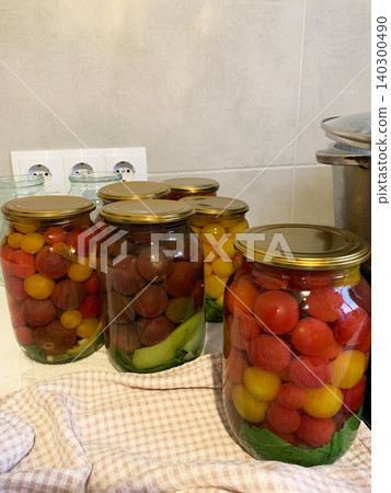 In a kitchen, jars filled with canned tomatoes sit on a table beside fresh vegetables. A mixing bowl holds whole tomatoes as preparation for winter storage continues In a kitchen, jars filled with canned tomatoes sit on a table beside fresh vegetables. A mixing bowl holds whole tomatoes as preparation for winter storage continues 140300490