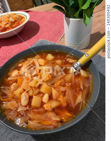 A bowl of Ukrainian borscht sits on a red cloth. The soup contains cabbage, carrots, and tomatoes. A silver spoon rests beside the bowl in a home kitchen space 140300492