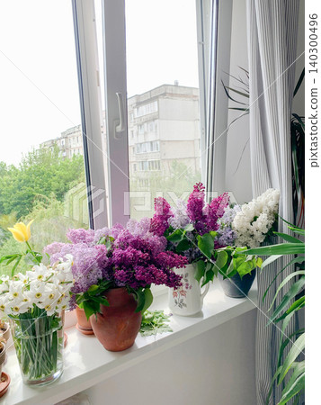 A vase with seasonal flowers sits on a windowsill in an apartment. Sunlight shines through the window, highlighting the colorful blooms and greenery 140300496