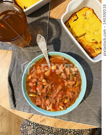 A bowl of Ukrainian borscht sits on a red cloth. The soup contains cabbage, carrots, and tomatoes. A silver spoon rests beside the bowl in a home kitchen space A bowl of Ukrainian borscht sits on a red cloth. The soup contains cabbage, carrots, and tomatoes. A silver spoon rests beside the bowl in a home kitchen space 140300538
