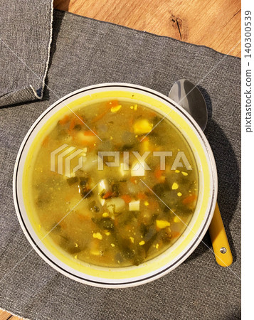 A bowl filled with potato and sorrel soup sits on a dark cloth. The soup has chunks of potatoes and bits of vegetables mixed in. A spoon rests beside the bowl A bowl filled with potato and sorrel soup sits on a dark cloth. The soup has chunks of potatoes and bits of vegetables mixed in. A spoon rests beside the bowl 140300539