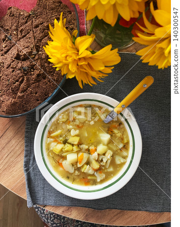 A bowl of soup containing potatoes, zucchini, and cauliflower sits on a wooden table. Fresh bread and yellow flowers are nearby, creating a simple meal scene A bowl of soup containing potatoes, zucchini, and cauliflower sits on a wooden table. Fresh bread and yellow flowers are nearby, creating a simple meal scene 140300547