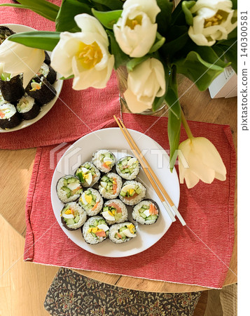 Sushi rolls are arranged neatly on a white plate. The rolls contain cucumber, red fish, and egg. They are served with chopsticks on a blue and green mat Sushi rolls are arranged neatly on a white plate. The rolls contain cucumber, red fish, and egg. They are served with chopsticks on a blue and green mat 140300581