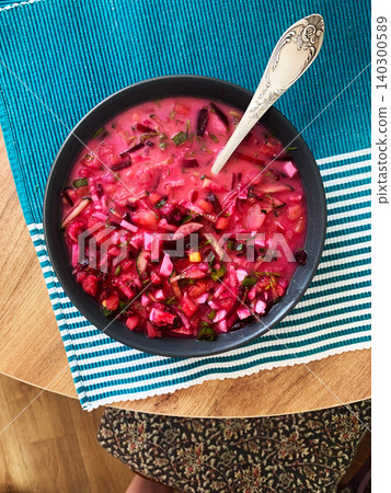 A bowl of cold beetroot soup sits on a blue and white tablecloth. Fresh vegetables and herbs are mixed into the soup. A silver spoon rests beside the bowl. The scene is set for lunch A bowl of cold beetroot soup sits on a blue and white tablecloth. Fresh vegetables and herbs are mixed into the soup. A silver spoon rests beside the bowl. The scene is set for lunch 140300589