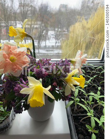 A vase with seasonal flowers sits on a windowsill in an apartment. Sunlight shines through the window, highlighting the colorful blooms and greenery 140300594