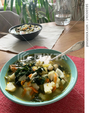 A bowl of nettle and egg soup is placed on a red cloth. The soup contains potatoes, cauliflower, and chopped greens. A spoon rests beside the bowl ready for use A bowl of nettle and egg soup is placed on a red cloth. The soup contains potatoes, cauliflower, and chopped greens. A spoon rests beside the bowl ready for use 140300595