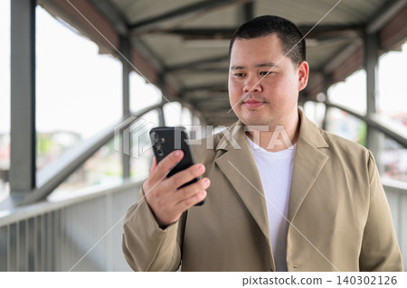 Plus size Isan Thai man looking down at phone on a bridge walkway Plus size Isan Thai man looking down at phone on a bridge walkway 140302126