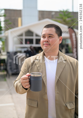 Plus size Isan Thai man holding a travel mug outdoors at a cafe Plus size Isan Thai man holding a travel mug outdoors at a cafe 140302138