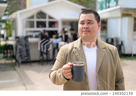 Plus size Isan Thai man holding a coffee mug outdoors at a cafe Plus size Isan Thai man holding a coffee mug outdoors at a cafe 140302139