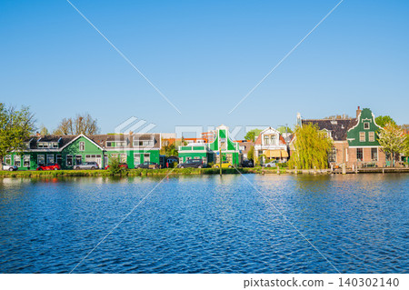 Amsterdam, Netherlands, 22.04.2026 - Traditional green wooden Dutch houses on the river bank in Zaanse Schans village with cars parked on the street Amsterdam, Netherlands, 22.04.2026 - Traditional green wooden Dutch houses on the river bank in Zaanse Schans village with cars parked on the street 140302140