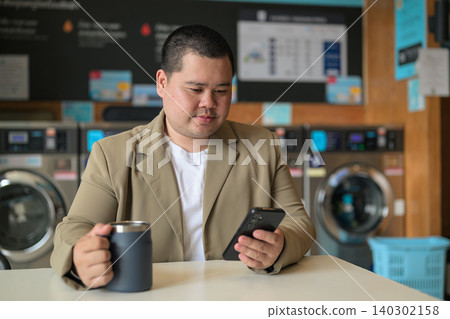 Plus size Isan Thai man using smartphone at a laundromat Plus size Isan Thai man using smartphone at a laundromat 140302158