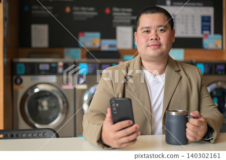 Plus size Isan Thai man looking at camera with phone and mug at a laundromat Plus size Isan Thai man looking at camera with phone and mug at a laundromat 140302161
