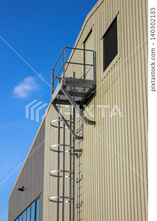 An industrial vertical fire escape ladder with a safety cage and landing platform is mounted on a beige corrugated metal wall. A ventilation louver and blue sky with light clouds are also visible. An industrial vertical fire escape ladder with a safety cage and landing platform is mounted on a beige corrugated metal wall. A ventilation louver and blue sky with light clouds are also visible. 140302185
