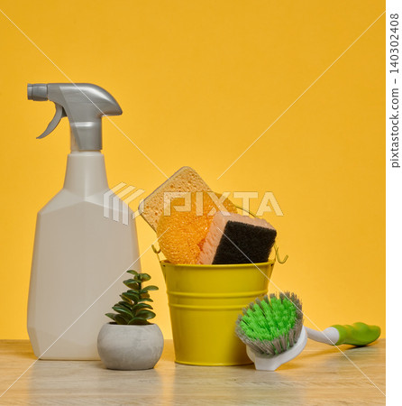 White plastic spray bottle and a stack of multi-colored sponges stand on a wooden surface against a yellow background. White plastic spray bottle and a stack of multi-colored sponges stand on a wooden surface against a yellow background. 140302408