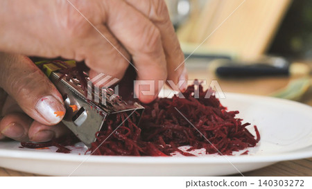 Hands grating fresh beetroot on a grater for healthy food preparation in a kitchen Hands grating fresh beetroot on a grater for healthy food preparation in a kitchen 140303272