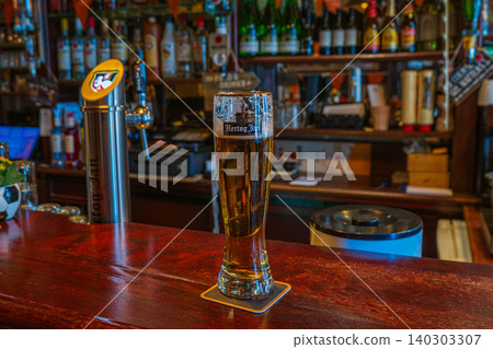 Amsterdam, Netherlands, 22.04.2026 - Hertog jan beer glass on wooden counter in traditional dutch bar interior, pint of draft lager on coaster with illuminated beer tap dispenser on background 140303307