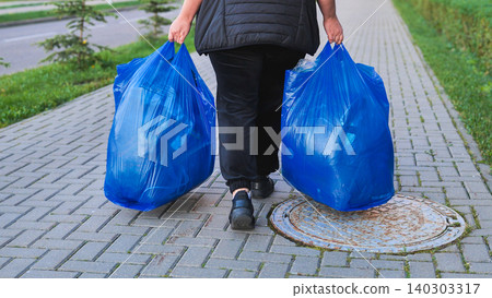 Person carrying blue trash bags on a paved sidewalk, walking towards disposal 140303317