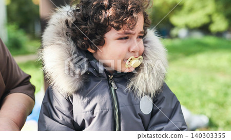 Toddler crying with pacifier, wearing winter jacket, experiencing sadness outdoors 140303355